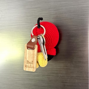 May include: A red crocheted keychain with a black stem and yellow accents, holding keys and a wooden tag. The tag is engraved with the words "Best Wishes". The keychain is attached to a silver ring.