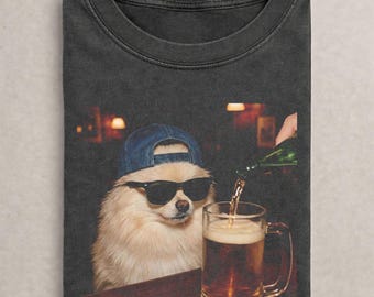 May include: Dark gray t-shirt featuring a Pomeranian dog wearing sunglasses and a blue baseball cap. The dog is sitting at a bar with a beer mug being filled. The background shows a bar setting.