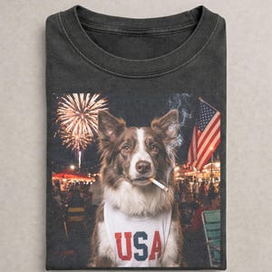 May include: A charcoal gray t-shirt featuring a graphic of a brown and white dog wearing a white tank top with the text USA printed in red and blue. The dog has a cigarette in its mouth with smoke rising, set against a background of fireworks.