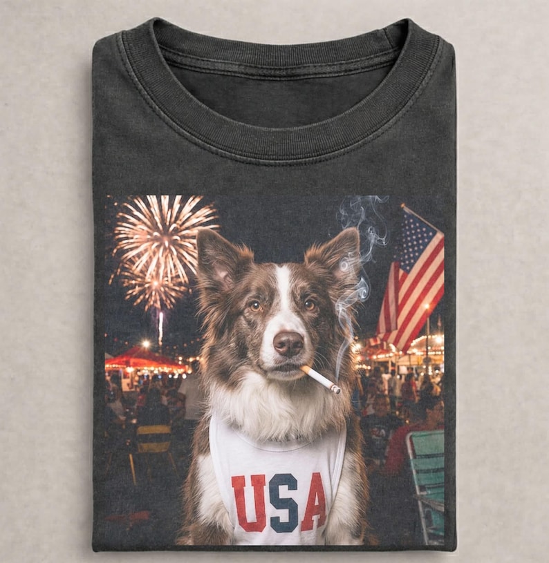 May include: A charcoal gray t-shirt featuring a graphic of a brown and white dog wearing a white tank top with the text USA printed in red and blue. The dog has a cigarette in its mouth with smoke rising, set against a background of fireworks.