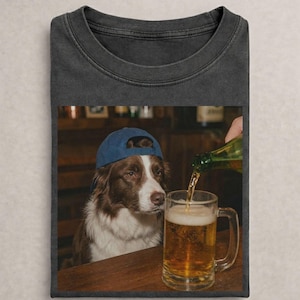 May include: A charcoal gray crew neck t-shirt featuring a graphic print of a brown and white dog wearing a blue baseball cap. The dog is sitting at a wooden table next to a glass beer mug being filled with a golden liquid from a green bottle.