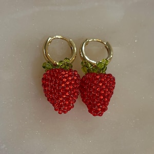 May include: A pair of gold hoop earrings with beaded strawberry charms. The strawberries are red with green beaded tops. The earrings are displayed on a light-colored surface.