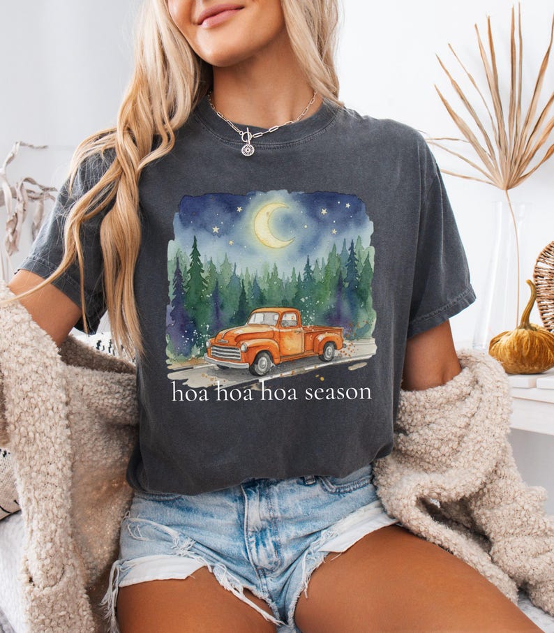 May include: Dark grey t-shirt with a watercolour design of an orange pickup truck on a road through a forest under a starry night sky with a crescent moon. The text "hoa hoa hoa season" is below the image.