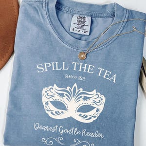 May include: A blue Comfort Colors t-shirt with the text "Spill the Tea since 1813" and a white masquerade mask design. A gold necklace with a small pendant rests on the shirt.