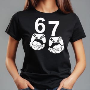 May include: Black t-shirt with the number 67 in white. Below the numbers are two white illustrations of video game controllers held by hands. The shirt is a casual style.