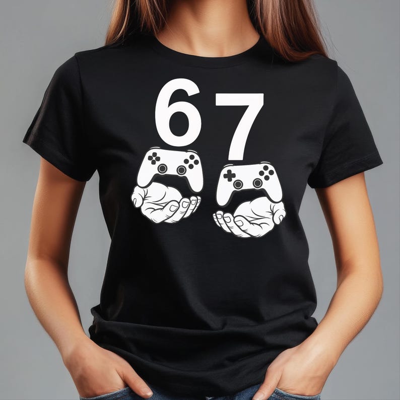 May include: Black t-shirt with the number 67 in white. Below the numbers are two white illustrations of video game controllers held by hands. The shirt is a casual style.