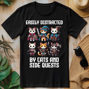 以下が含まれることがあります： 「EASILY DISTRACTED BY CATS AND SIDE QUESTS」の白文字がプリントされた黒いTシャツ。ファンタジー衣装を着た7匹の漫画の猫のグラフィック。鎧、マント、帽子など。デザインは赤、オレンジ、青を使用しています。