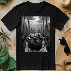 Może przedstawiać: Czarny t-shirt z szarą grafiką mopsa z dużymi oczami, na tle lasu z trzema postaciami kosmitów. Projekt jest wyśrodkowany na koszulce, tworząc uderzający kontrast wizualny.