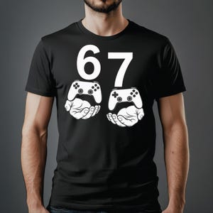 May include: Black t-shirt with a white graphic. The graphic features the number "67" above two white hands holding white video game controllers. The design is centered on the chest.