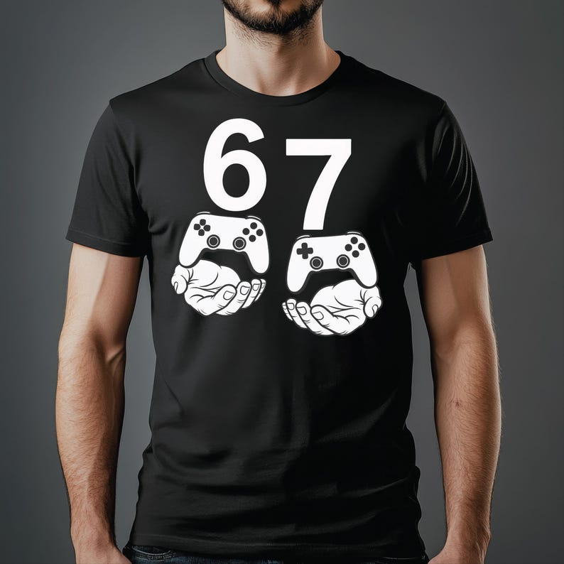 May include: Black t-shirt with a white graphic. The graphic features the number "67" above two white hands holding white video game controllers. The design is centered on the chest.