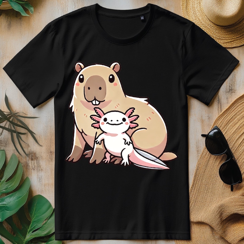 Pode incluir: Uma camiseta preta com uma imagem de desenho animado de uma capivara e uma axolotl. A capivara &eacute; marrom e tem o nariz rosa. A axolotl &eacute; branca com br&acirc;nquias rosas.