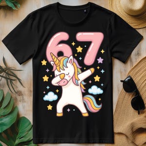 May include: Black t-shirt with a unicorn dabbing, featuring a rainbow mane and tail. The number "67" is in pink above the unicorn, surrounded by stars and clouds. A fun birthday shirt.