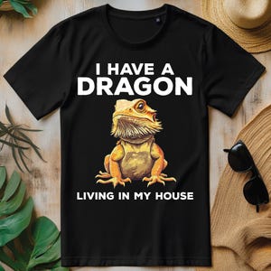 Pode incluir: Camiseta preta com o texto branco "I HAVE A DRAGON LIVING IN MY HOUSE". A camiseta apresenta uma ilustração detalhada de um dragão barbudo em tons de amarelo, laranja e marrom.