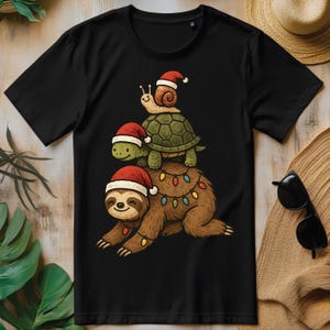 May include: Black t-shirt featuring a whimsical Christmas design. A sloth, turtle, and snail are stacked, each wearing a Santa hat. The sloth is adorned with Christmas lights. The design is a fun, festive graphic.