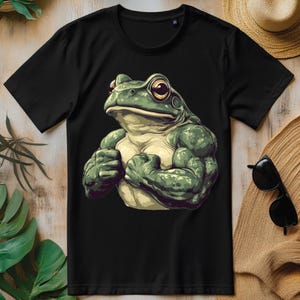 Może przedstawiać: Czarny t-shirt z zieloną, kreskówkową żabą o wybulonych mięśniach. Żaba zgina bicepsy i ma poważny wyraz twarzy.