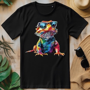 Könnte beinhalten: Schwarzes T-Shirt mit einer farbenfrohen Illustration einer Bartagame mit Sonnenbrille. Die Eidechse ist in leuchtenden Farben von Rot, Blau, Gelb und Grün dargestellt und bildet einen auffälligen visuellen Kontrast zum dunklen Hintergrund.