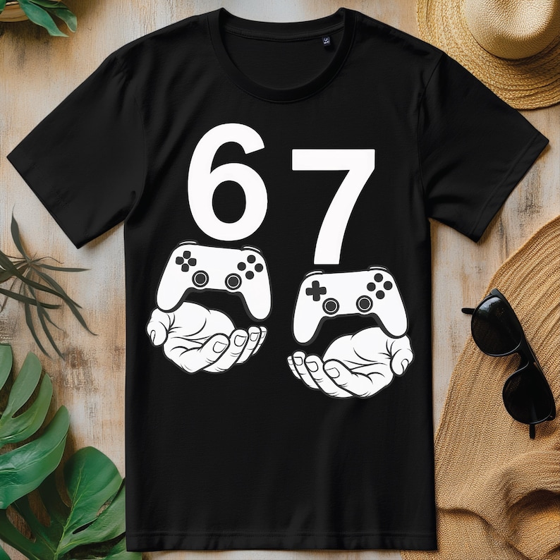 May include: Black t-shirt with the number 67 in white. Below the numbers are two white illustrations of hands holding video game controllers. The controllers have black buttons and directional pads.