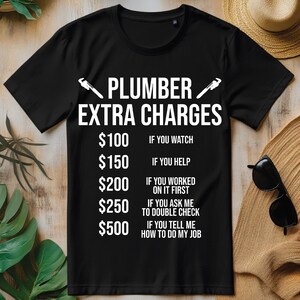Puede incluir: Camiseta negra con texto blanco que dice "PLUMBER EXTRA CHARGES" y una lista de precios y razones para cargos adicionales. Los precios son 100 €, 150 €, 200 €, 250 € y 500 €.