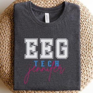 May include: Dark grey heather t-shirt featuring the text "EEG TECH Jennifer" in white, blue, and pink. The shirt is made of a soft material and is displayed on a woven, circular surface.