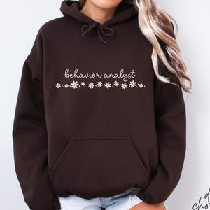Op de afbeelding: Donkerchocoladebruine hoodie met een voorzak en een capuchon met trekkoord. De woorden "behavior analyst" zijn in cursief geschreven, met daaronder een rij kleine madeliefjes. De woorden "dark chocolate" zijn in script rechtsonder geschreven.