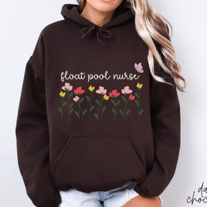 Könnte beinhalten: Dunkelbrauner Kapuzenpullover mit Fronttasche und Kordelzugkapuze. Auf der Vorderseite steht der Text "float pool nurse" über einer Reihe bunter Blumen und einer Schmetterlingsgrafik. Die Worte "dark chocolate" sind unten rechts in Kursivschrift geschrieben.