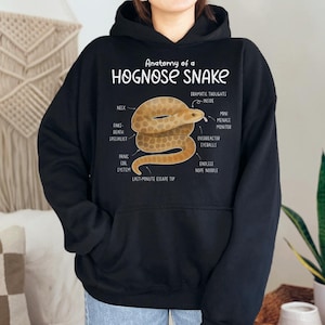 Könnte beinhalten: Schwarzer Kapuzenpullover mit einer Grafik einer Hakennasennatter und anatomischen Beschriftungen. Der Text "Anatomy of a Hognose Snake" befindet sich über der Schlangenillustration. Der Hoodie hat eine Kängurutasche und eine Kapuze mit Kordelzug.