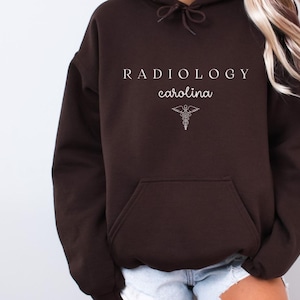 Può includere: Una felpa con cappuccio marrone scuro con il testo "RADIOLOGY carolina" e un simbolo medico bianco.