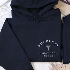 Peut inclure: Sweat-shirt à capuche noir avec le texte "SCARLETT FLOAT POOL NURSE" en blanc. Le sweat-shirt a une capuche à cordon et des poignets côtelés. Le design comprend un symbole médical.