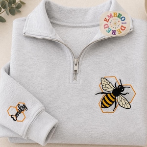 Peut inclure: Un sweat-shirt gris clair à fermeture éclair quart de longueur avec une abeille brodée sur des contours hexagonaux orange sur la poitrine. La manche affiche le nom Emily en écriture noire dans un petit hexagone orange. Un logo circulaire indique EMBROIDERED.