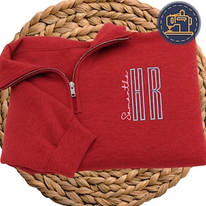 May include: A red quarter-zip sweatshirt with the name "Samantha" embroidered in cursive, and the letters "HR" in a light blue, block-style font. The sweatshirt is displayed on a woven, circular surface. A small, circular patch with a sewing machine is in the upper right corner.