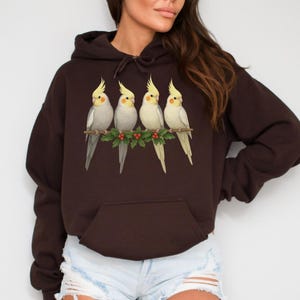 Peut inclure: Un sweat à capuche marron foncé avec un motif de quatre cacatoès perchés sur une branche avec du houx. Les oiseaux ont des crêtes jaunes et des plumes blanches et grises. Le sweat a une poche avant et une capuche à cordon.