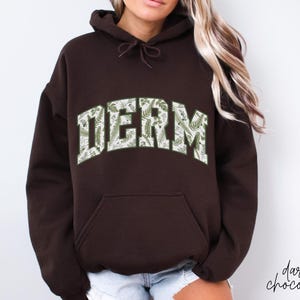 Pu&ograve; includere: Una felpa con cappuccio marrone scuro con una stampa floreale bianca e verde che scrive "DERM".