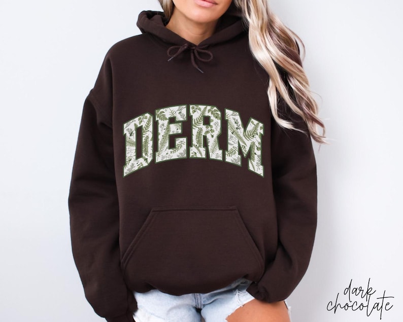 Pu&ograve; includere: Una felpa con cappuccio marrone scuro con una stampa floreale bianca e verde che scrive "DERM".