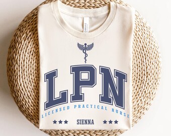 May include: A natural-colored t-shirt with the text "LPN" in navy blue collegiate lettering, above the words "LICENSED PRACTICAL NURSE" and the name "SIENNA". A navy blue caduceus symbol is above the "LPN" text. The word "natural" is in the bottom right corner.