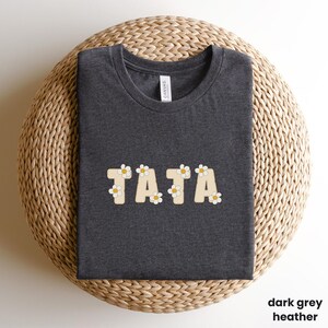 May include: Dark grey heather t-shirt with the word "TATA" in a cream-colored, daisy-decorated font. The shirt is displayed on a woven, light brown surface. The text "dark grey heather" is in the bottom right corner.