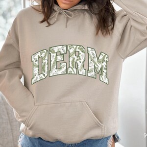 Pu&ograve; includere: Una felpa con cappuccio beige con una grande stampa floreale della parola "DERM" in un font in stile universitario.