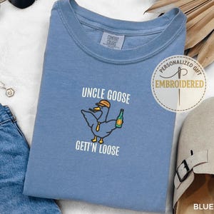 以下が含まれることがあります： 「UNCLE GOOSE GETT'N LOOSE」の刺繍と、帽子とボトルを持った漫画のアヒルのイラストが描かれた、ブルーデニムカラーのTシャツ。クルーネックで、アクセサリーに囲まれています。