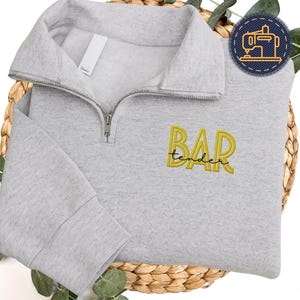 May include: A pale grey quarter-zip sweatshirt with the words "BAR tender" embroidered in yellow. The sweatshirt features a high collar and a silver zip. It is displayed on a woven basket.