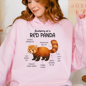 May include: Pink hoodie with a graphic of a red panda anatomy. The illustration includes labels such as "suspicious cinnamon fox," "marshmallow ring tail," and "sleep deprived eyes." The text "Anatomy of a Red Panda" is at the top.