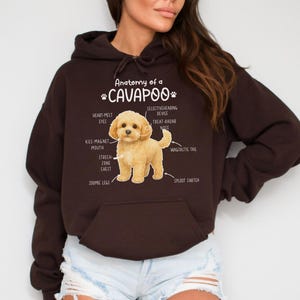 Könnte beinhalten: Ein dunkelbrauner Kapuzenpullover mit einer Grafik eines Cavapoo-Hundes und seiner beschrifteten Anatomie. Der Text lautet "Anatomie eines Cavapoo" mit Bezeichnungen wie "Herzschmelz-Augen" und "Wackeliger Schwanz". Der Hoodie hat eine Vordertasche und eine Kapuze mit Kordelzug.