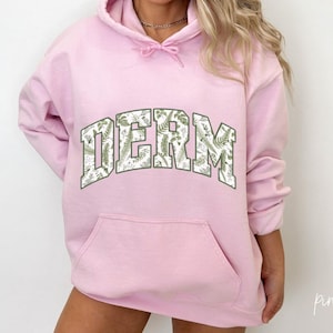 Pu&ograve; includere: Felpa rosa con stampa floreale bianca e la parola "DERM" in un grande carattere grassetto.