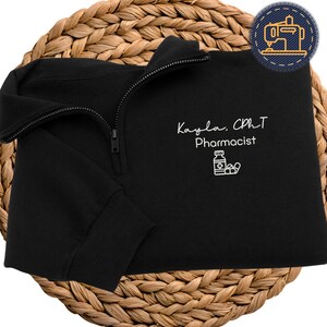 Puede incluir: Sudadera negra con cremallera de cuarto de cremallera con el nombre "Kayla, CPhT Pharmacist" y un gráfico de botella de pastillas en blanco. La sudadera tiene una cremallera negra y mangas largas. Un parche circular con una máquina de coser está en la esquina superior derecha.