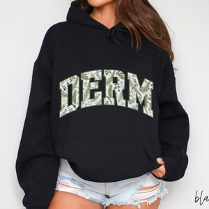 Pu&ograve; includere: Felpa con cappuccio nera con una stampa floreale bianca e verde che dice "DERM" sul davanti.