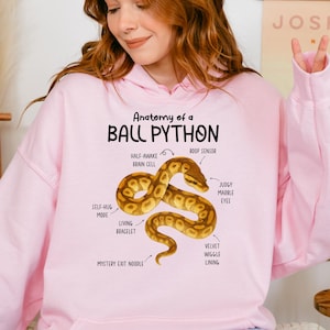 Peut inclure: Un sweat à capuche rose clair avec un motif d'un python royal et son anatomie. Le design comprend des étiquettes telles que "Half-Awake Brain Cell" et "Judgy Marble Eyes". Le texte en haut indique "Anatomy of a Ball Python".