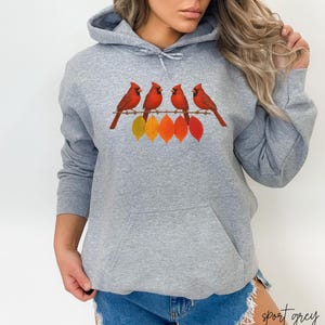 Könnte beinhalten: Ein hellgrauer Kapuzenpullover mit einer Vordertasche und einem Kordelzug. Er zeigt eine Grafik von vier roten Kardinälen auf einem Ast mit bunten Blättern. Der Text "sport grey" ist unten rechts aufgedruckt.