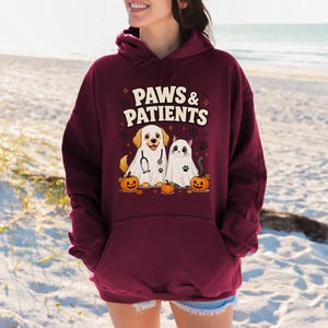 May include: A maroon hooded sweatshirt featuring a graphic print with the text PAWS & PATIENTS. The design shows a dog and a cat dressed as ghosts, each wearing a white sheet, accompanied by three orange jack-o'-lantern pumpkins and paw prints.