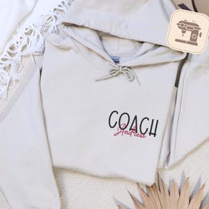 May include: A beige hoodie with a drawstring hood and the word "COACH" in black script, with "Andrew" in pink script underneath. The hoodie is made of a soft material and is likely for casual wear.