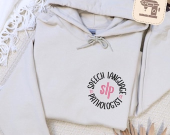 Op de afbeelding: Een beige hoodie met een trekkoord en een buidelzak. De voorkant heeft een cirkelvormig ontwerp met de tekst "SPEECH LANGUAGE PATHOLOGIST" en de initialen "SLP" in roze.