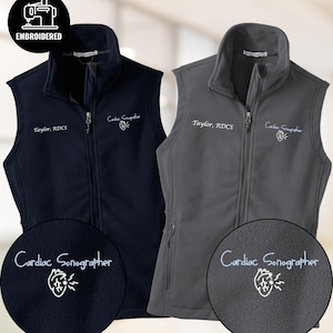 Könnte beinhalten: Zwei Fleece-Westen, eine marineblaue und eine graue, mit Reißverschluss vorne und hohem Kragen. Jede Weste hat die gestickten Texte "Taylor, RDCS" und "Cardiac Sonographer" mit einer Herzgrafik. Das Wort "EMBROIDERED" befindet sich in einem Kreis.