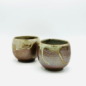 May include: Two brown ceramic cups with a white glaze. The glaze has a drip effect, creating a unique pattern on each cup.